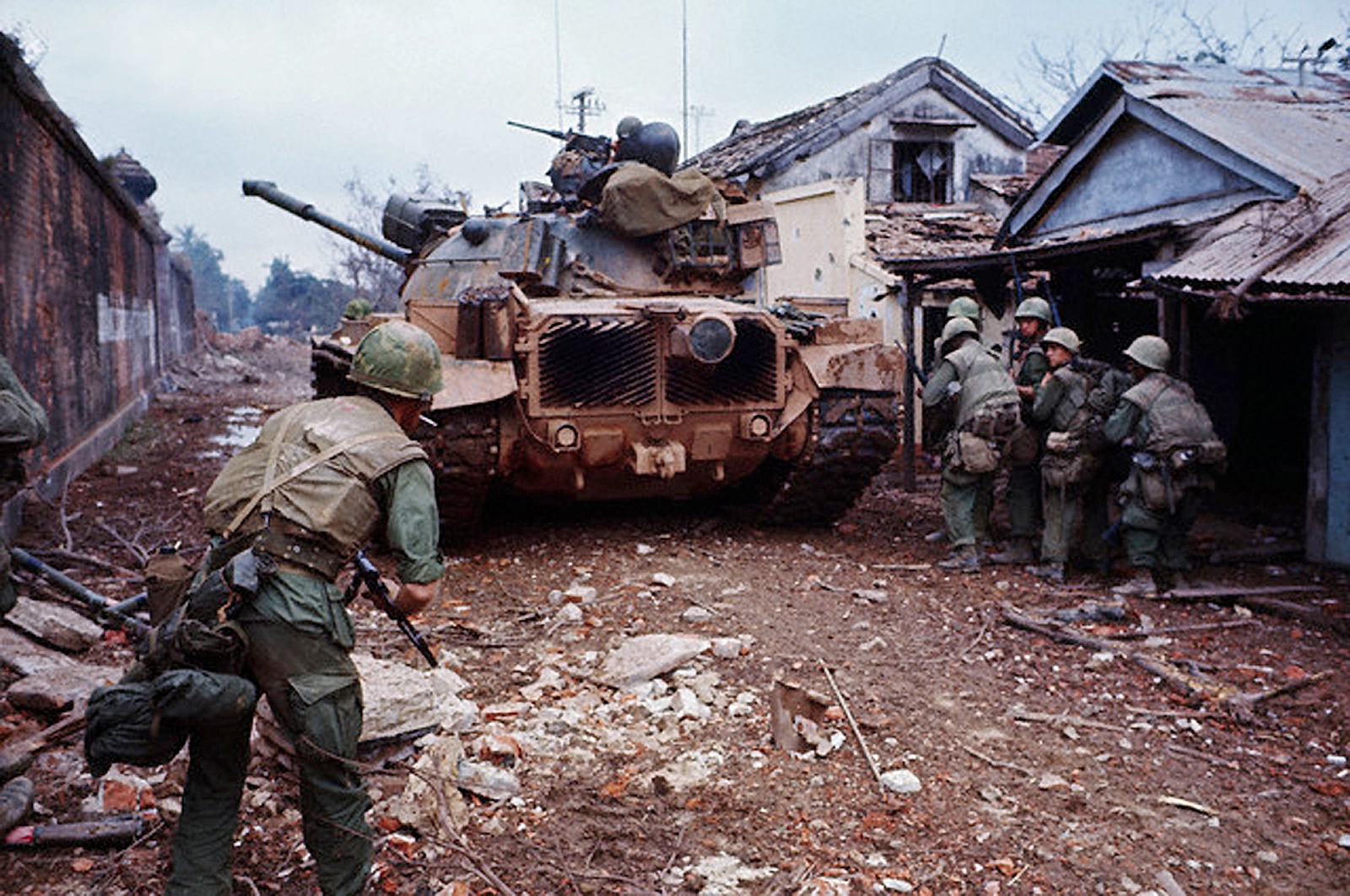Vietnam War Engagement, Combat Ready Soldier, M48 Patton Tank, Urban Warfare, Military Readiness, Tactical Positioning, Armored Vehicles, M16 Rifle, Helmeted Soldier, Mud-Covered Tank, Devastated Urban Area, Battle Aftermath, Warzone Debris, Soldiers in Combat, War Tactics, Armed Conflict, Military Operation, Soldier Vigilance, Infantry Tactics, Color War Photography