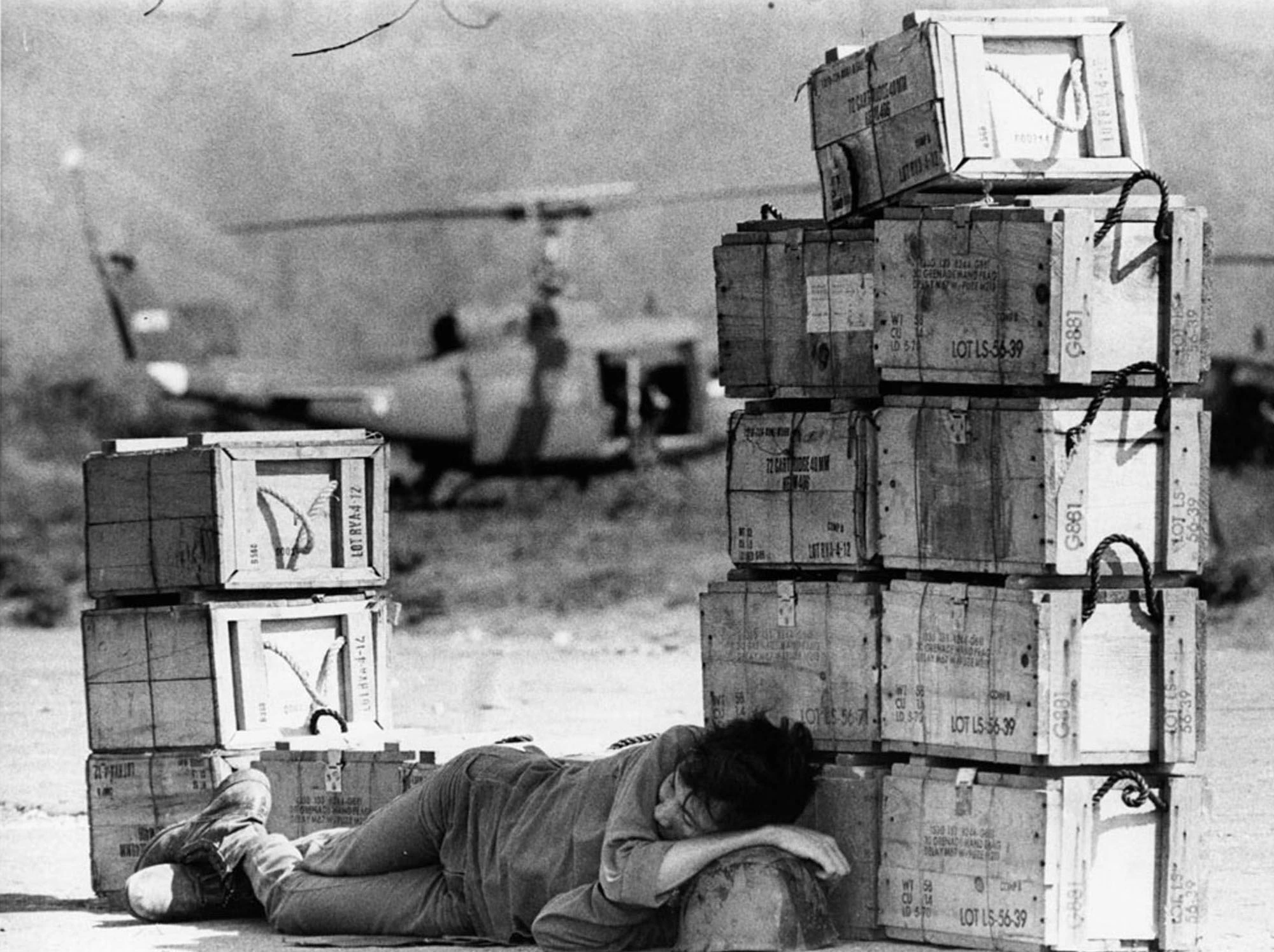 Resting Soldier, Ammunition Boxes, Black and White, Military Helicopter, Military Base, Landing Zone, Soldier Fatigue, War Conditions, Resupply Operation, Field Operations, War Photography, Soldier's Vulnerability, Moment of Rest, Combat Environment, Historical Military, Military Logistics, Warzone Fatigue, Soldier Respite, Conflict Snapshot